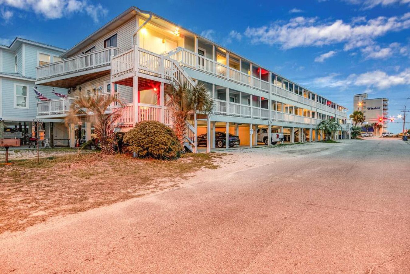 A multi-story hotel with a balcony on the ground floor, a parking area in front, and a clear sky.