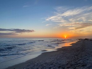 Une plage où le soleil se couche à l’horizon, les vagues venant doucement lécher le rivage, et quelques personnes dispersées le long de la plage.