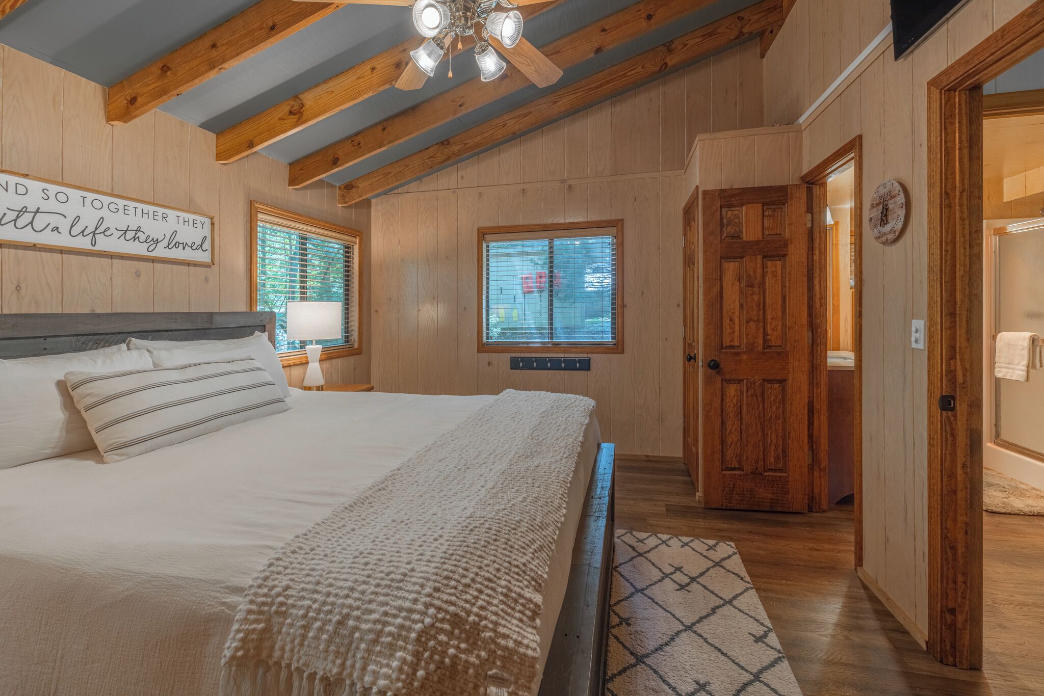 Een slaapkamer met een houten plafond, een bed met witte lakens, een houten deur en een wandklok.