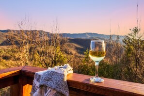 Un bicchiere di vino bianco su una ringhiera di legno, con una coperta a maglia accanto, con vista su un paesaggio montuoso.