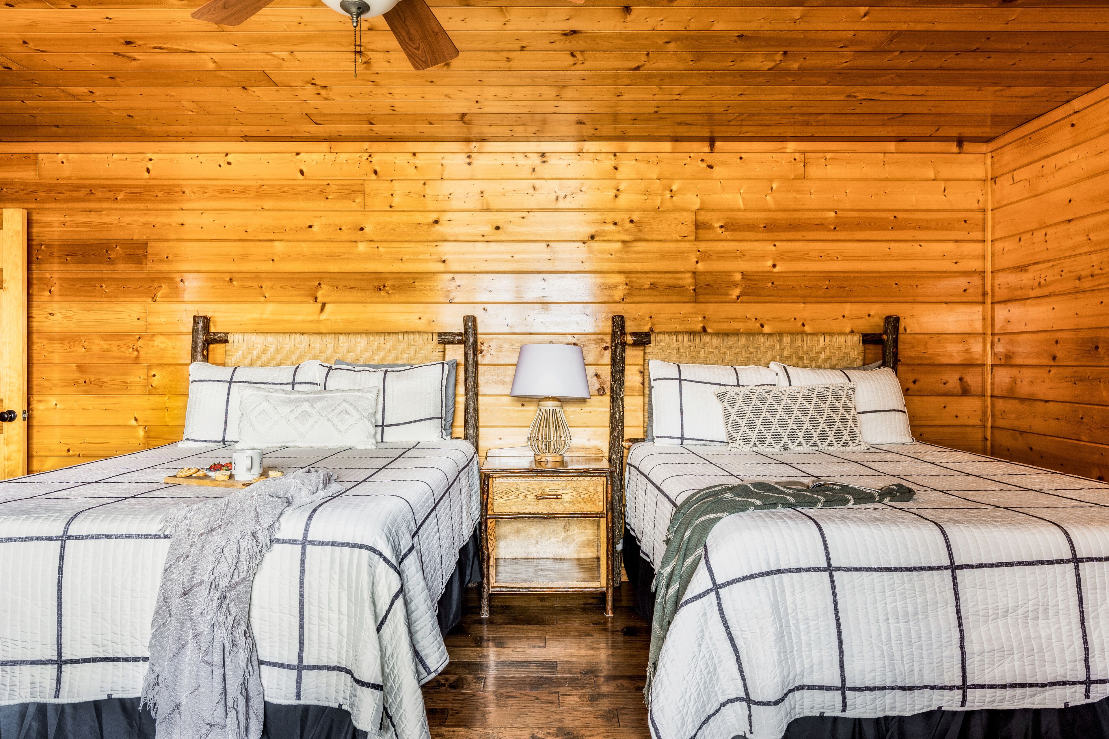 A wooden cabin room with two beds, a nightstand with a lamp, and a wooden ceiling fan.