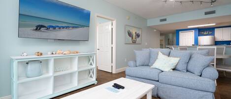 A living room with a large TV, a white coffee table, a blue sofa, and a ceiling fan.