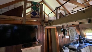 A loft-style room with a large TV, wooden beams, a hanging quilt, and a kitchen area with a sink and cabinets. - Near SkiSawmill 5 mi ETAR,STAR Archery, PAWA events. On the stream!   (Waterville)