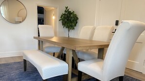 A dining area with a wooden table, four upholstered chairs, a bench, a mirror, a potted plant, and a wall-mounted light fixture. - Your Family Friendly Home with Large Backyard & Onsite Parking (New Rochelle)