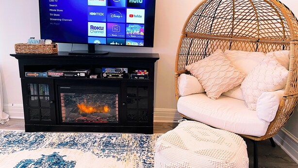 A living room with a flat-screen TV displaying a streaming service interface, a fireplace, a wicker chair, and a patterned rug. - Your Family Friendly Home with Large Backyard & Onsite Parking (New Rochelle)