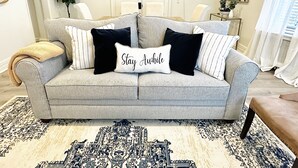A living room with a grey sofa, patterned rug, and a 'Stay Awake' pillow. - Your Family Friendly Home with Large Backyard & Onsite Parking (New Rochelle)