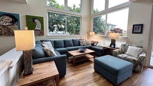 Amplia sala de estar con sofá modular, una mesa de centro y un sillón estampado. Amplias ventanas ofrecen luz natural y vistas a la vegetación exterior.