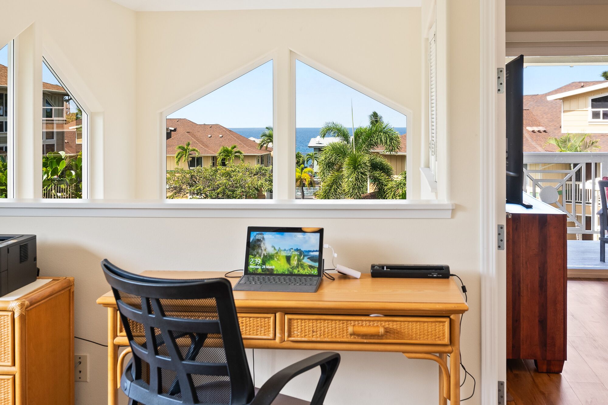 Ein Zimmer mit Schreibtisch, Laptop und Stuhl. Ein Fenster bietet Blick auf Häuser und Palmen.