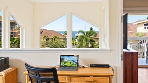 Habitación con escritorio, laptop y silla. Se ve una ventana con vista a casas y palmeras.