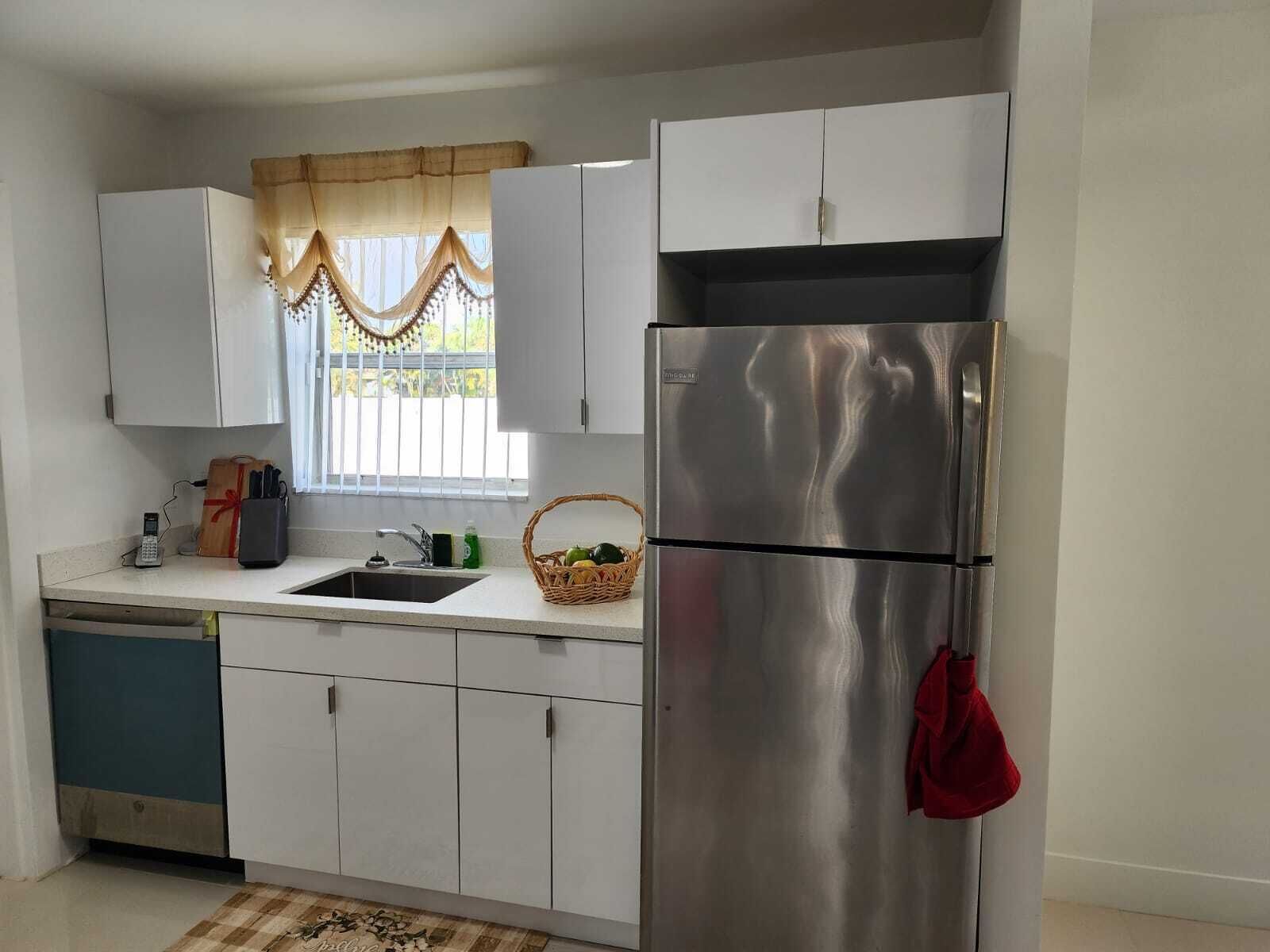 A compact kitchen with white cabinets, a stainless steel refrigerator, a sink, and a window with a curtain.