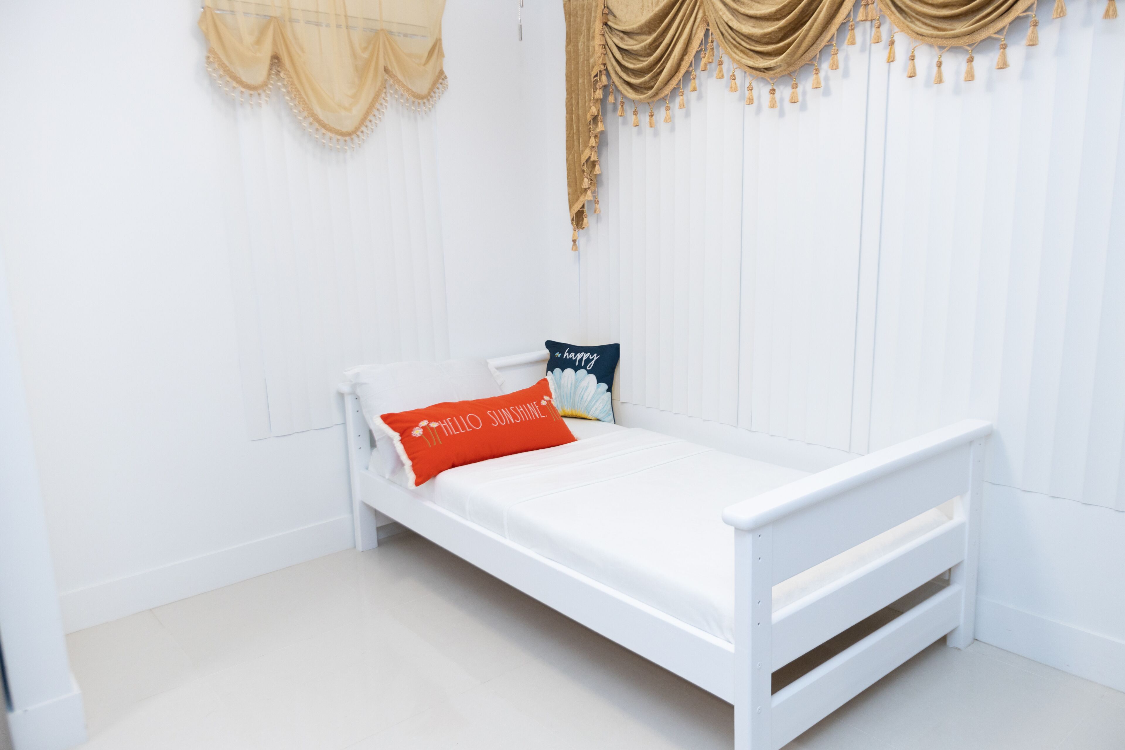Un lit simple blanc, avec un matelas blanc et deux oreillers, un rouge et un bleu, dans une pièce aux murs et au sol blancs.
