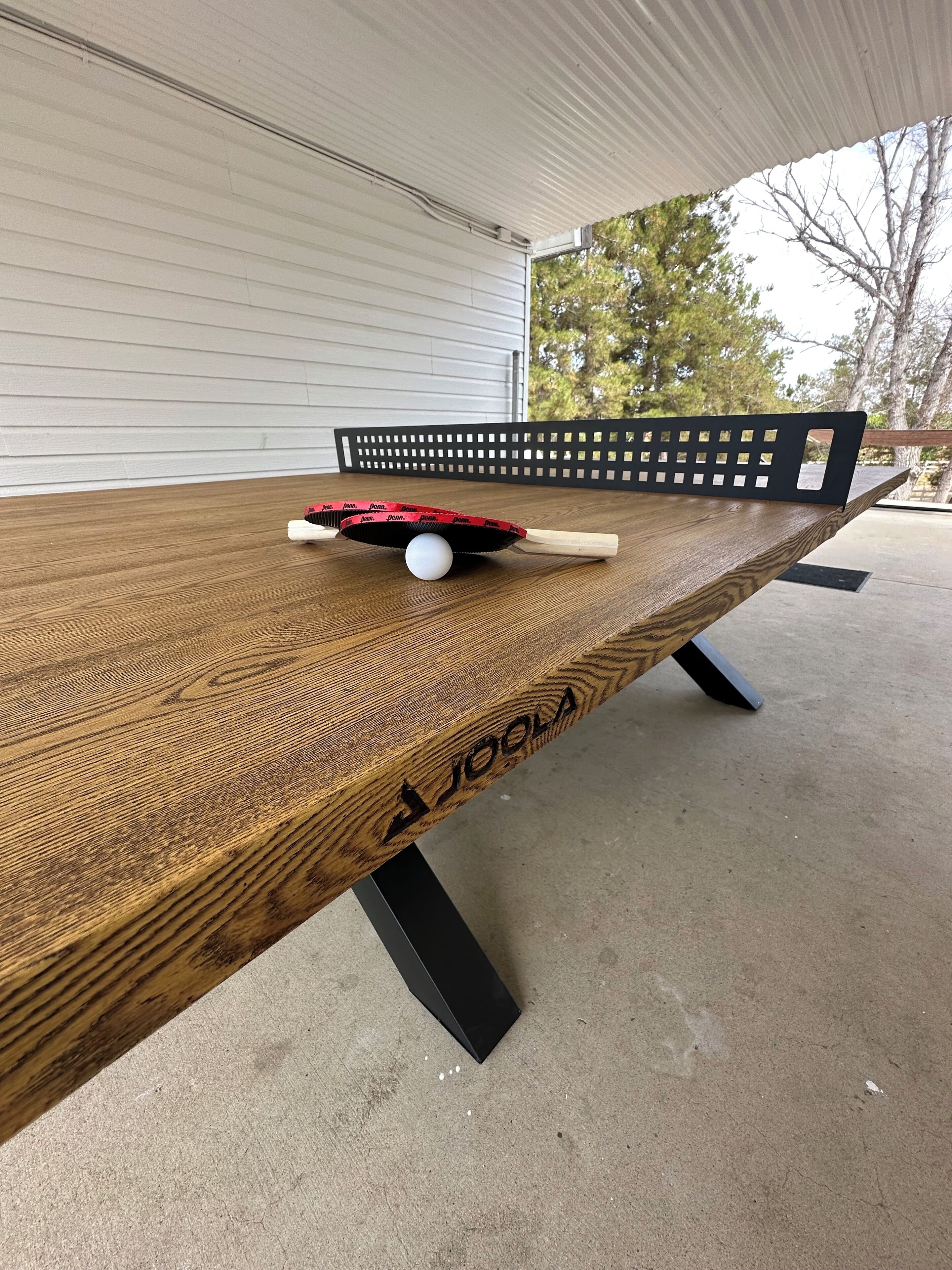 A wooden table with a white ball and a paddle, set outdoors with a metal bench and trees in the background.