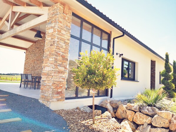 Una casa moderna con un pilar de piedra, ventanas amplias y un patio con sillas y un pequeño árbol.
