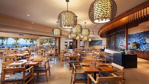 Un vaste espace dédié à la salle à manger, avec des tables et des chaises en bois, de grandes suspensions lumineuses et une vue sur l’océan.