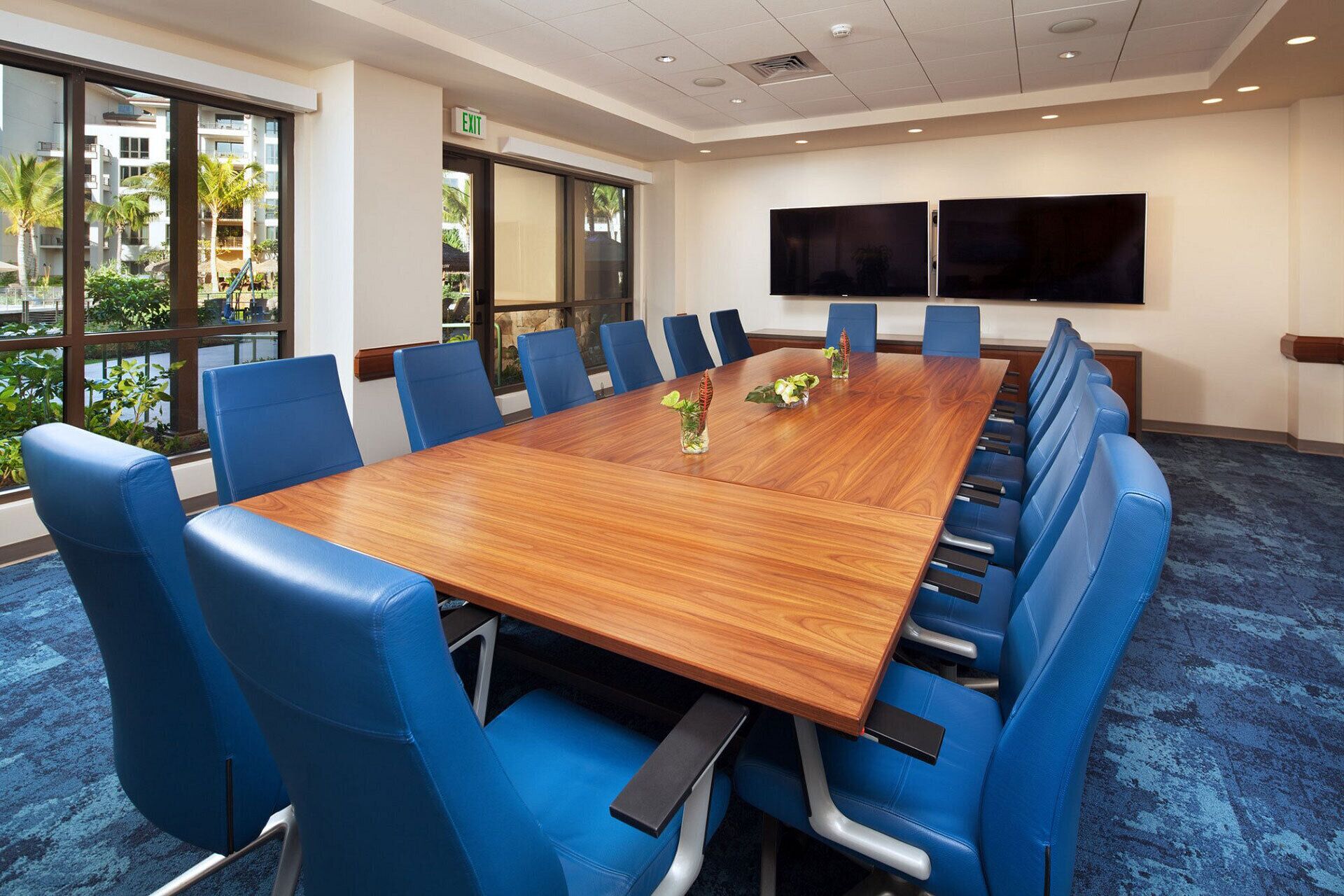 Une salle de conférence avec une longue table en bois, des chaises bleues et deux grands téléviseurs à écran plat fixés au mur.