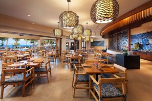 A spacious restaurant with wooden tables and chairs, large hanging lamps, and a view of the ocean.