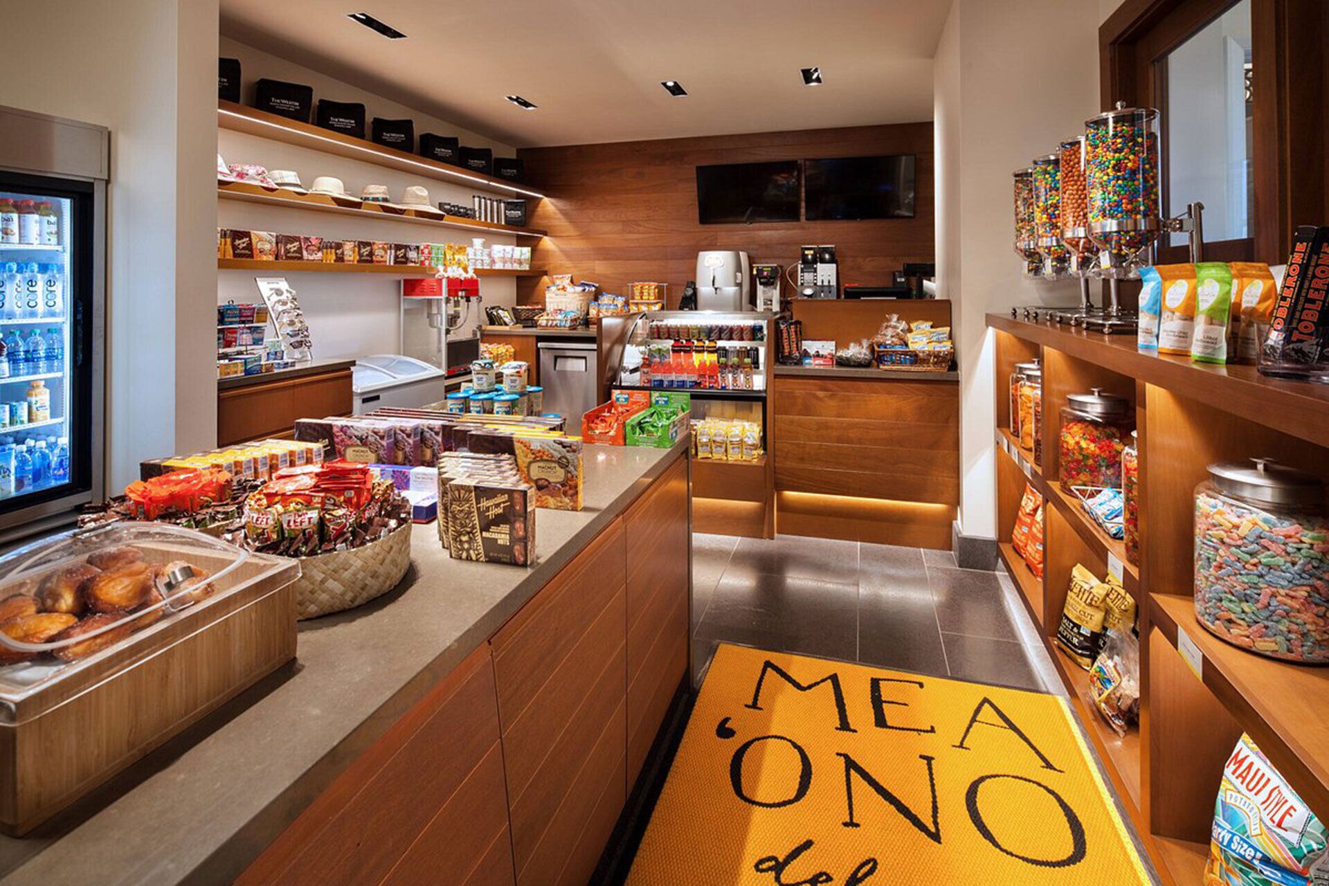 Um saguão de hotel com diversas opções de comida e bebida, incluindo uma máquina de café, um refrigerador com água engarrafada e uma seleção de lanches e acompanhamentos.