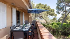An outdoor dining area with a table set for four, a blue umbrella, and a view of trees and the sea. - Marysol by Interhome (Les Issambres)