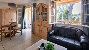 A living room with a black leather couch, wooden cabinet, dining table with chairs, and a window with a view of trees. - Marysol by Interhome (Les Issambres)