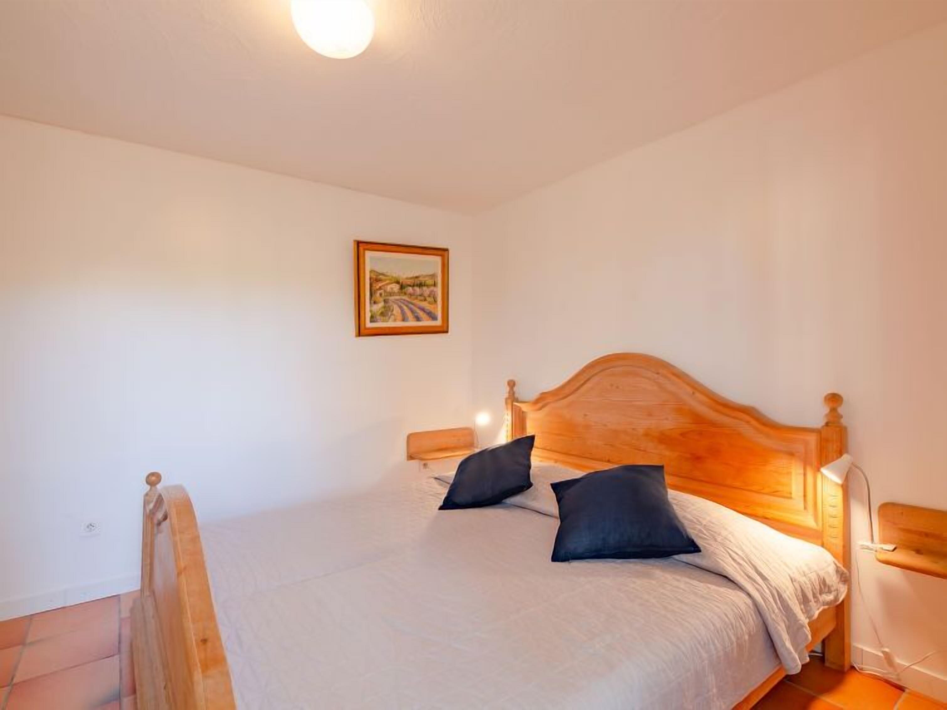 Un dormitorio con cama de madera, dos almohadas, una mesita de noche, una lámpara de pared y un cuadro en la pared.