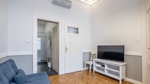 Un soggiorno con un divano blu, una televisione su un mobile bianco e una porta che dà su un corridoio.