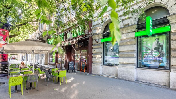 Un caffè all'aperto con tavoli e sedie, un ombrellone con marchio Coca-Cola e un edificio con segnaletica verde.