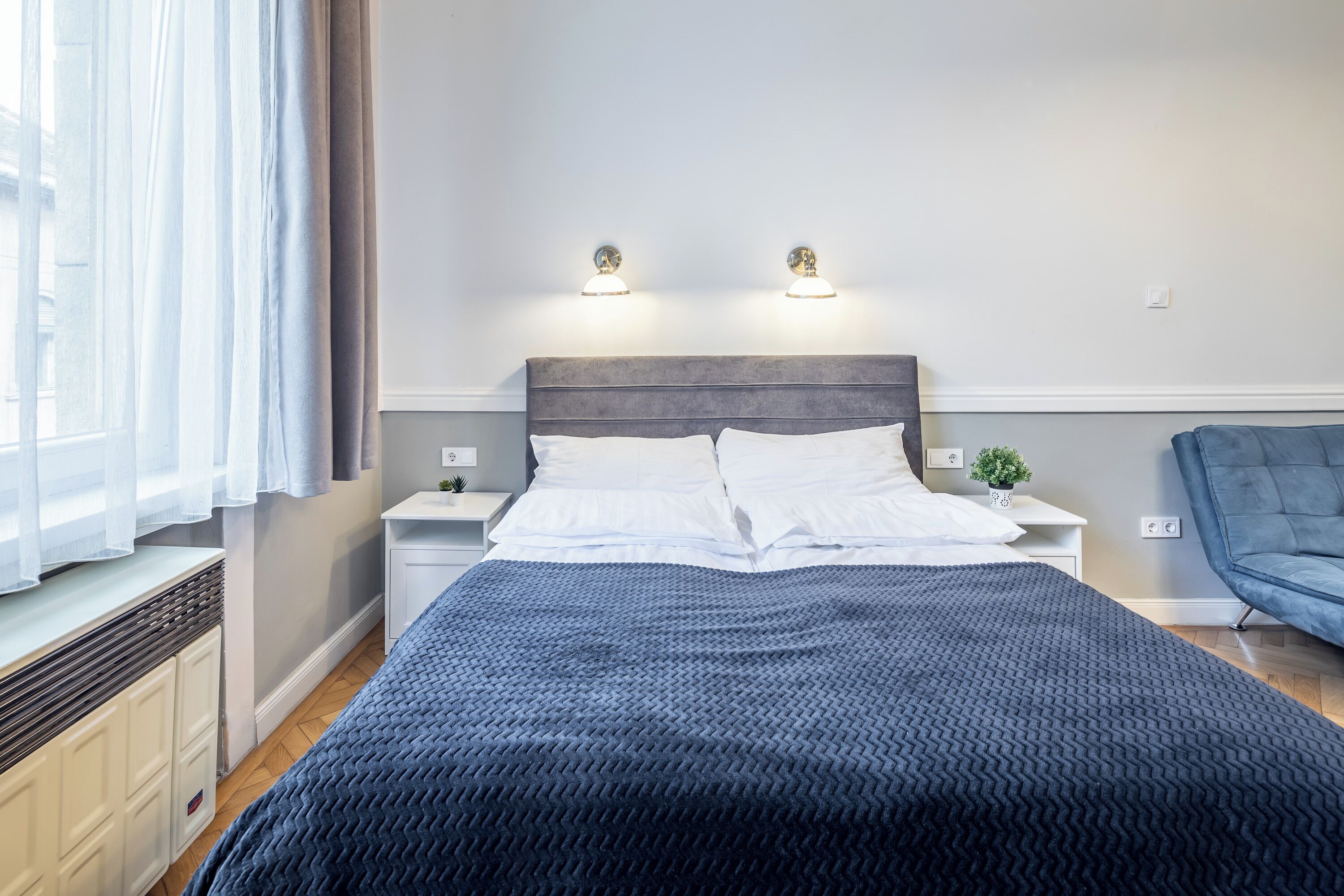 A neatly made bed with white linens, a dark blue textured blanket, and two bedside lamps.