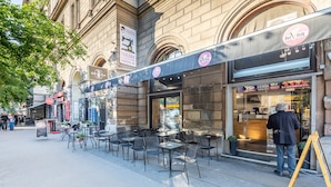 An outdoor café with tables and chairs, a sign for a tailor shop, and a restaurant with a menu board. - Central Luxury Apartment (Budapest)