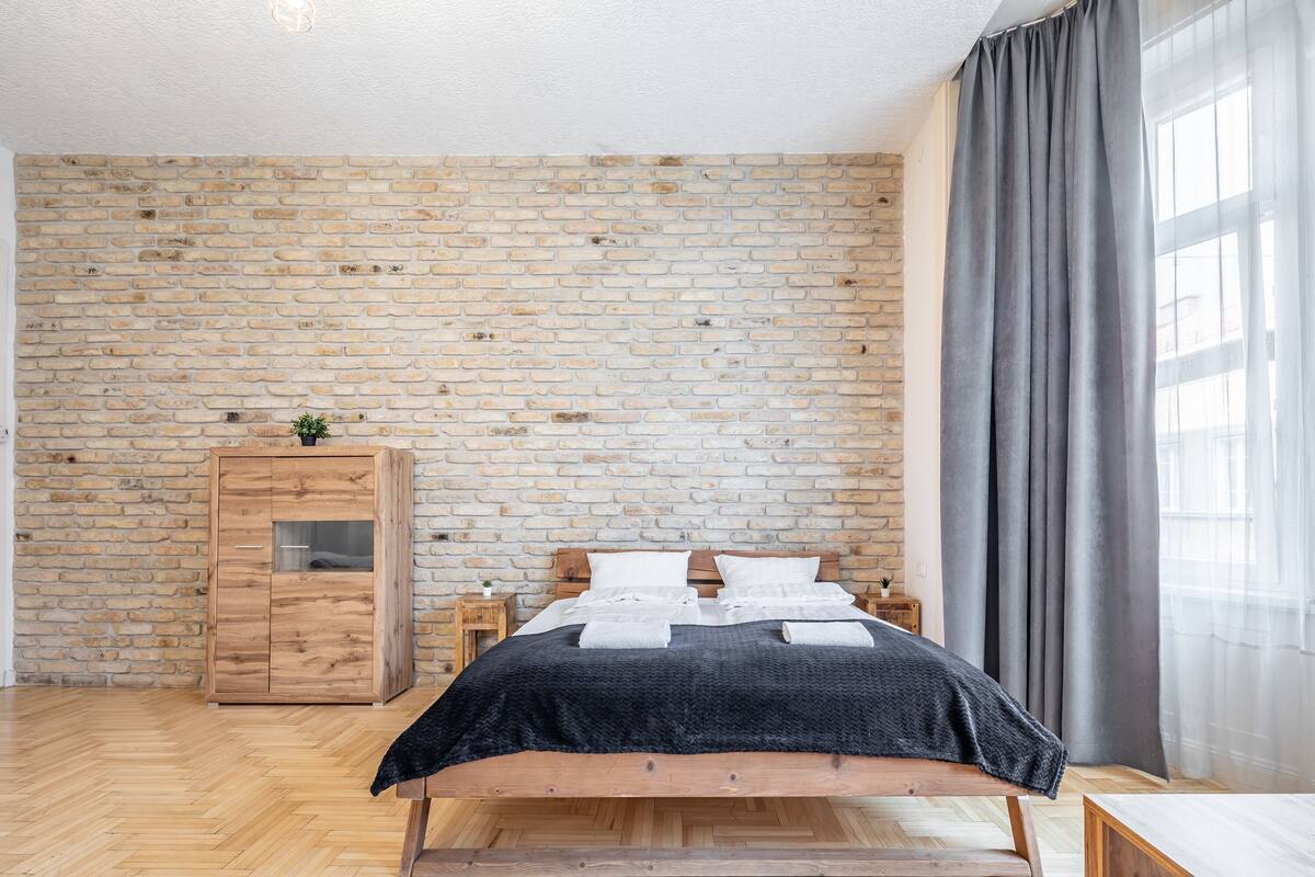 A bedroom with a brick wall, a wooden bed, a wardrobe, and a window with curtains.