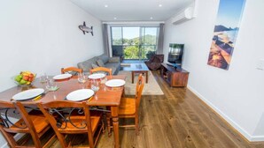 A dining area with a wooden table set for four, a sofa, and a TV in the background. - Shore Thing - Shoal Bay (Shoal Bay)