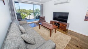 A living room with a sofa, a coffee table, a TV, and a balcony with a view of greenery. - Shore Thing - Shoal Bay (Shoal Bay)