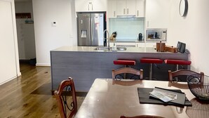 A modern kitchen with a central island, stainless steel appliances, and a dining area with a wooden table and chairs. - Shore Thing - Shoal Bay (Shoal Bay)
