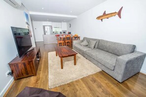 Ein modernes Wohnzimmer mit einem grauen Sofa, einem Holztisch und einem Fernseher auf einem Holzständer. Im Hintergrund ist eine Küche mit einem Essbereich zu sehen.