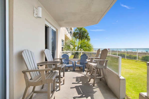 A balcony with wooden chairs and tables, a view of the ocean, and a grassy area.