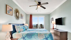 Un dormitorio con cama, dos lámparas de noche, ventilador de techo, televisor y vistas al exterior.