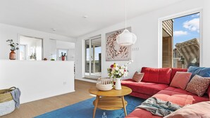 Een moderne woonkamer met een rode bank, een blauw vloerkleed en een houten salontafel. Er hangt een witte hanglamp en een schilderij aan de muur.