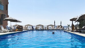Una piscina con sillones de descanso y sombrillas, rodeada por un edificio con vistas despejadas al mar.