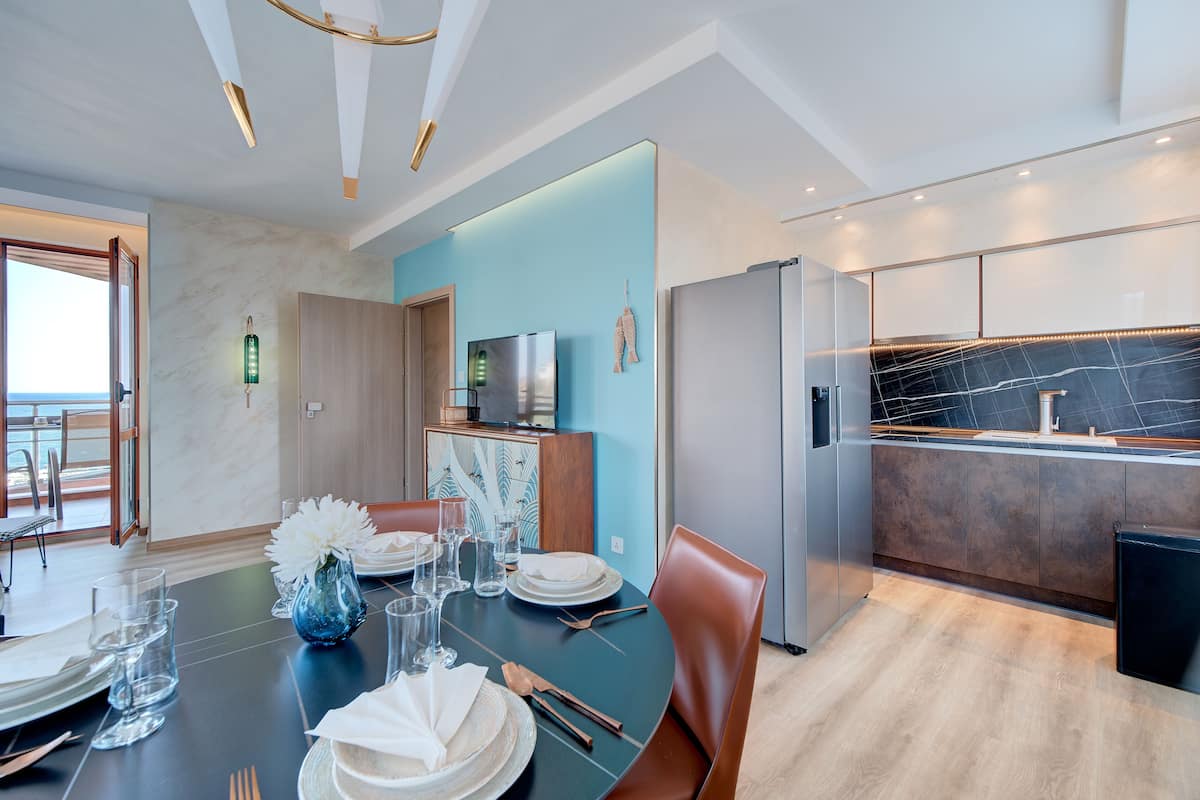 Un comedor moderno con una mesa redonda dispuesta para cuatro personas, una cocina con encimeras de mármol oscuro y vista al mar a través de un ventanal grande.