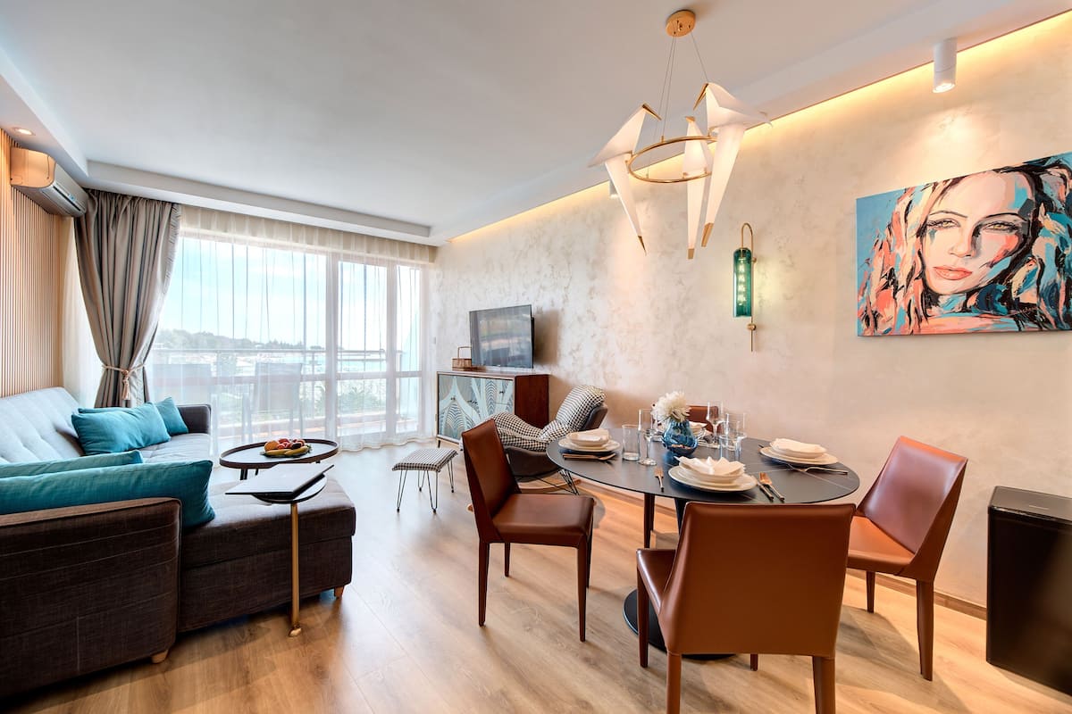 Una sala moderna con sofá, mesa de comedor y sillas. Hay un televisor, un cuadro en la pared y una ventana con cortinas.