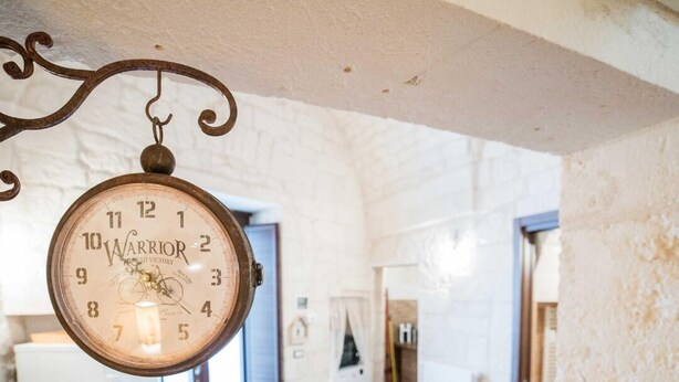 Ein geräumiges Zimmer mit einer großen, aufwendigen Uhr, die von der Decke hängt, einem Essbereich mit Tisch und Stühlen sowie einem Waschraum im Hintergrund.