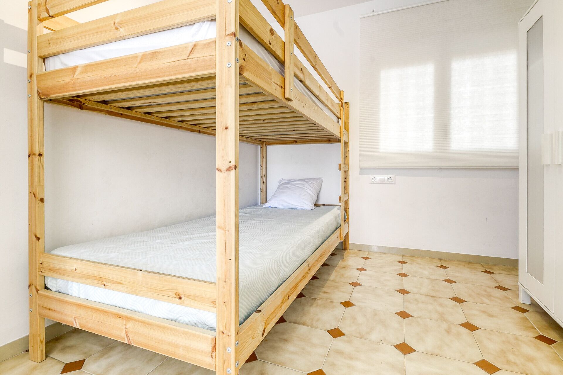 A bunk bed with a white mattress and a wooden frame in a room with a tiled floor and a white door.