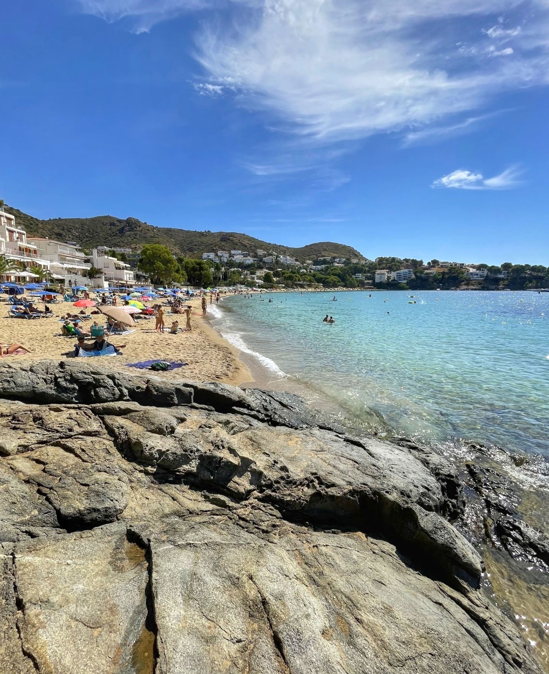A beach with clear water, rocky foreground, and buildings in the background.