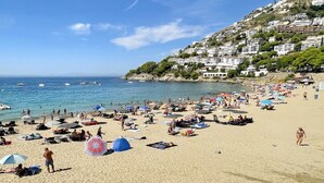 Ein Strand mit Menschen, die sonnenbaden und schwimmen, einem klaren, blauen Himmel und Gebäuden, die an einem Hügel im Hintergrund liegen.
