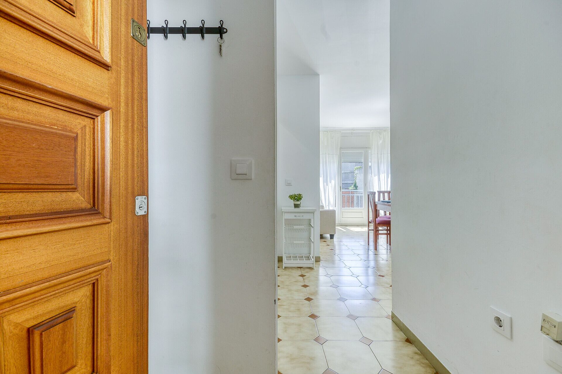 Ein Flur mit einer Holztür, einer weißen Wand, einem Fliesenboden, einem kleinen weißen Schrank mit Pflanze und Blick in einen Raum mit Tisch und Stühlen.