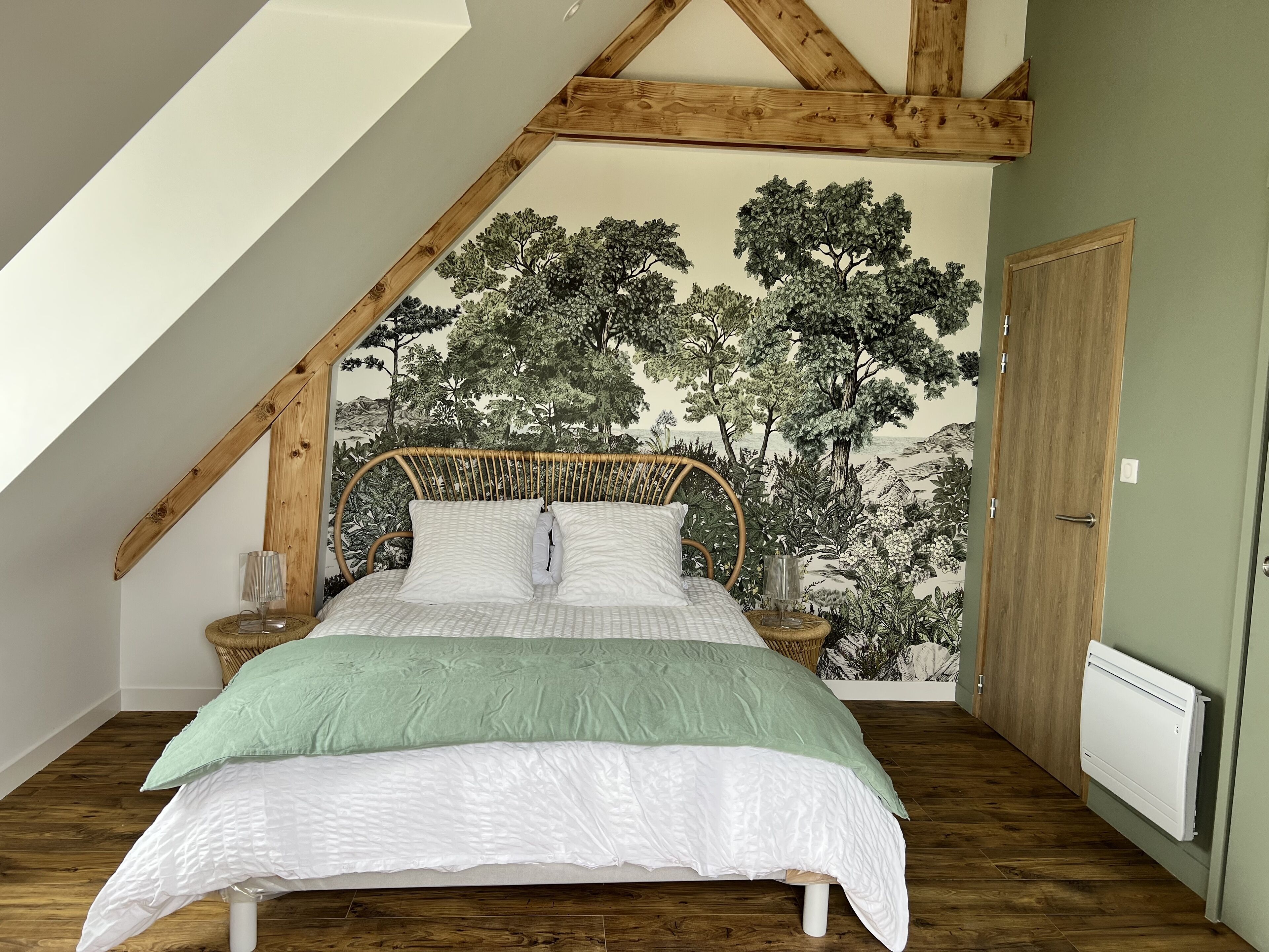 Een slaapkamer met een hoog plafond, houten balken, een bed met een groen-witte dekbedovertrek, een rieten hoofdbord en een muurschildering van bomen op de muur.