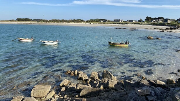A beach with boats anchored in calm waters, clear skies, and a rocky shoreline.
