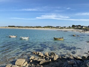 A beach with boats anchored in calm waters, clear skies, and a rocky shoreline.