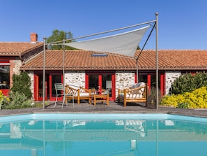A poolside area with a covered seating area, a stone building with red accents, and a clear blue pool. - Spacious family lodge with natural pool, near Nantes (La Limouzinière)
