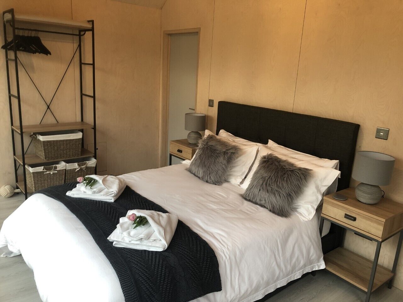A neatly made bed with white linens, two fluffy pillows, and a black throw blanket. There are two bedside tables with lamps and a small shelf with baskets.