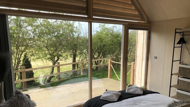 Ein Schlafzimmer mit großem Fenster, einem Bett, einem Sessel und einer Holzdielen-Terrasse.